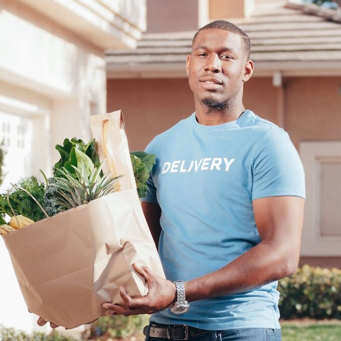 Smiling delivery man holding a paper bag of groceries on a sunny day outdoors.